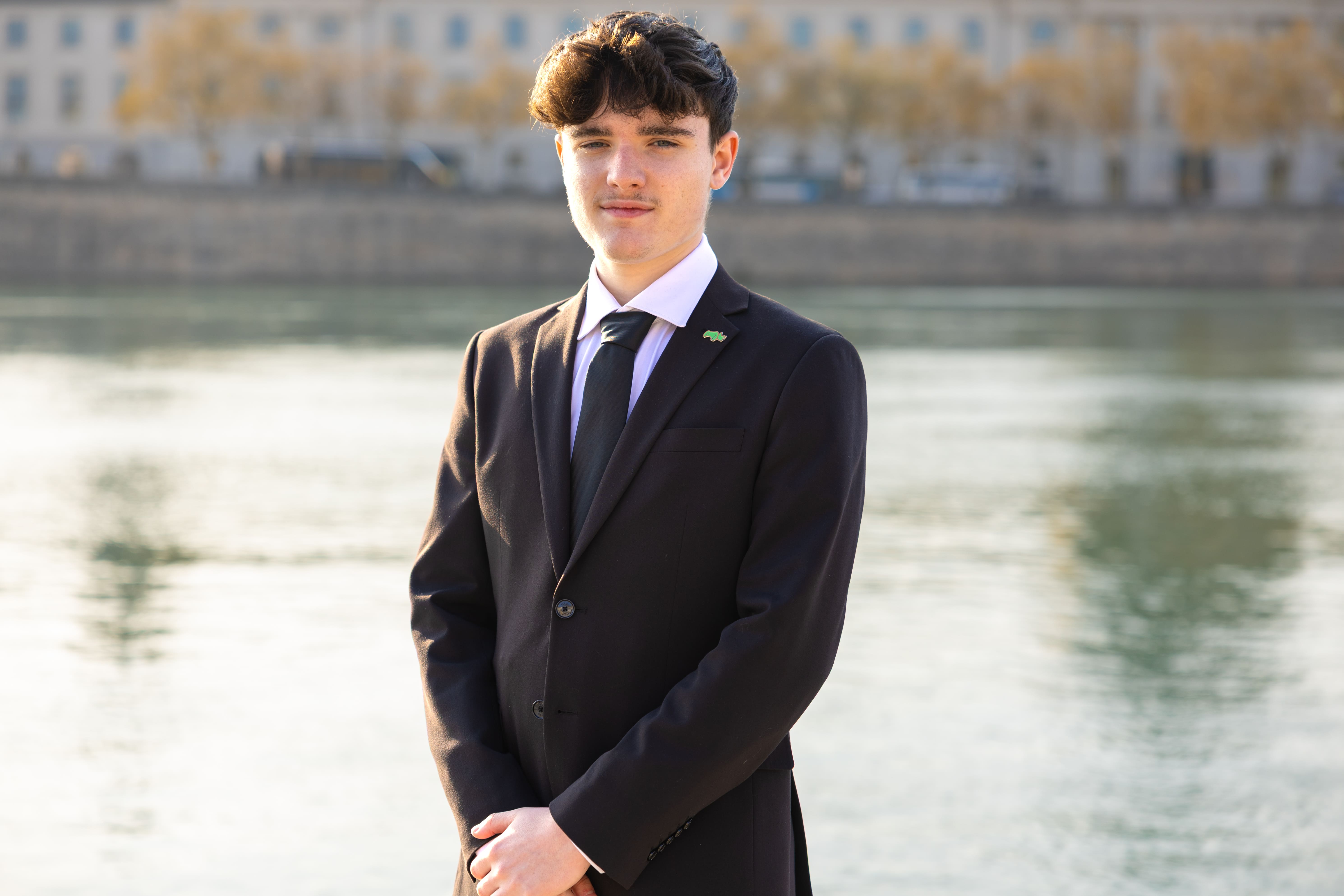 Young man with dark hair wearing a black suit and tie standing outdoors near a body of water with buildings in the background.