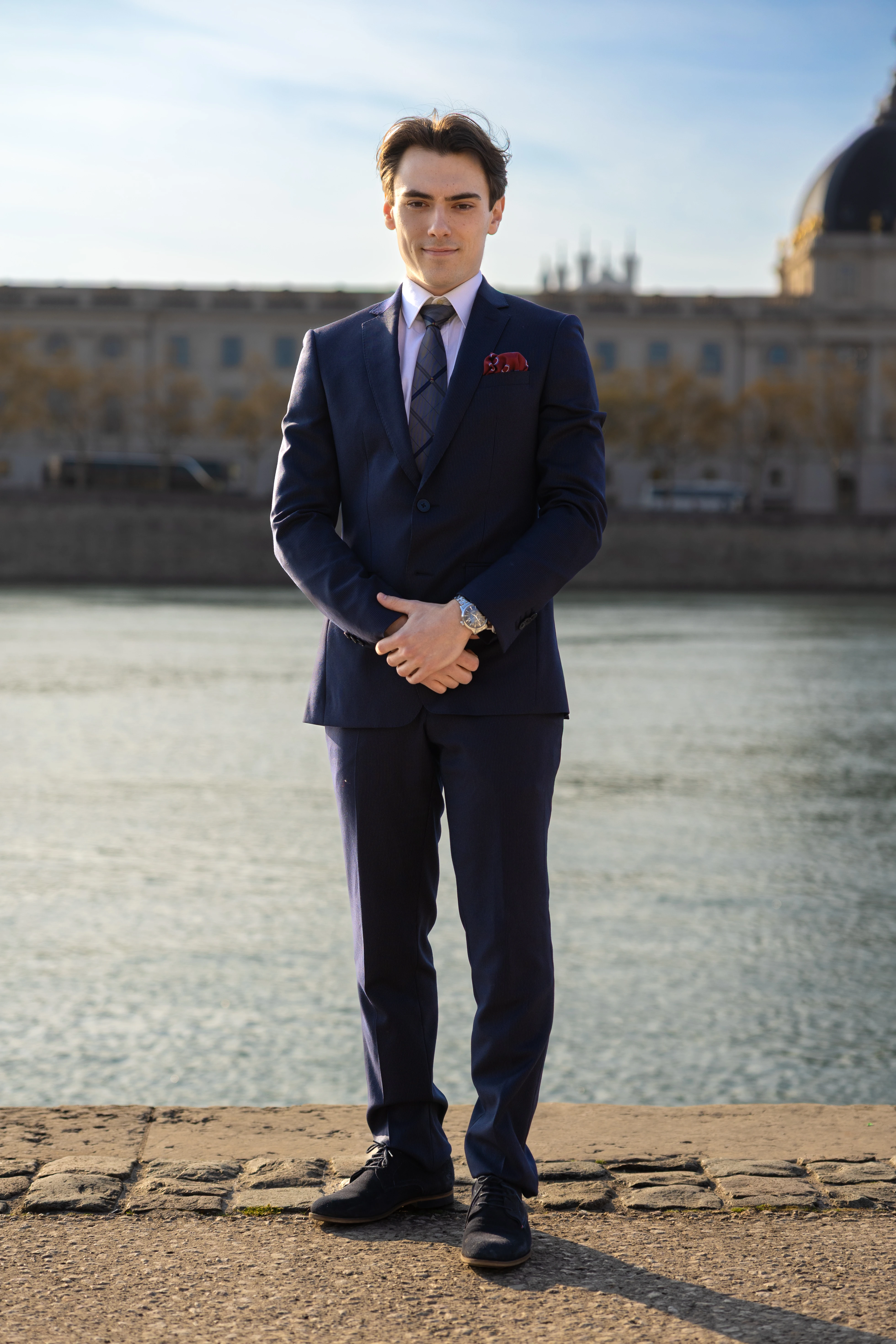 Young man in a navy suit with a patterned tie standing in front of a river with a blurred building in the background.