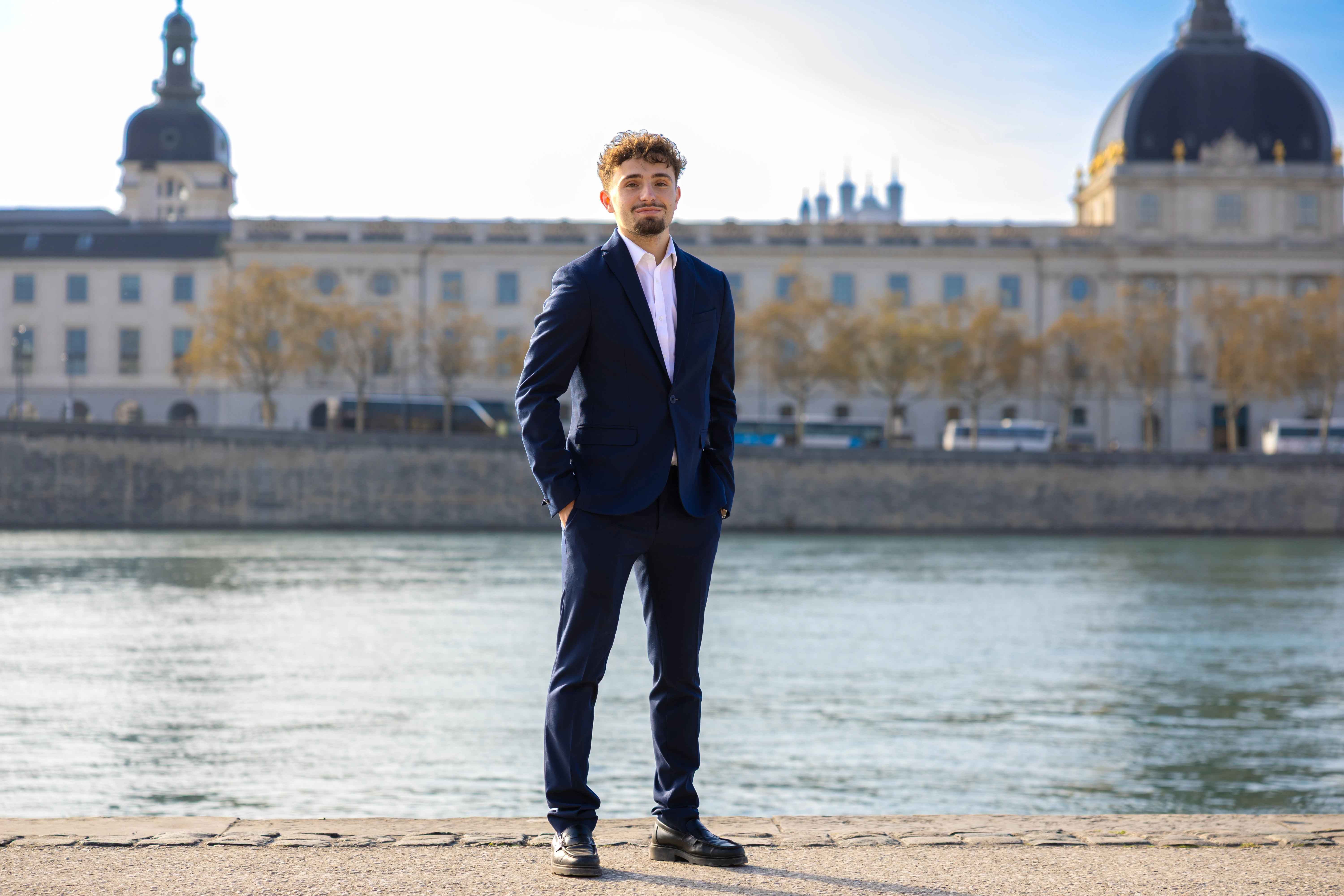 Young man with curly hair and beard wearing a navy blue suit and white shirt standing by a waterfront.