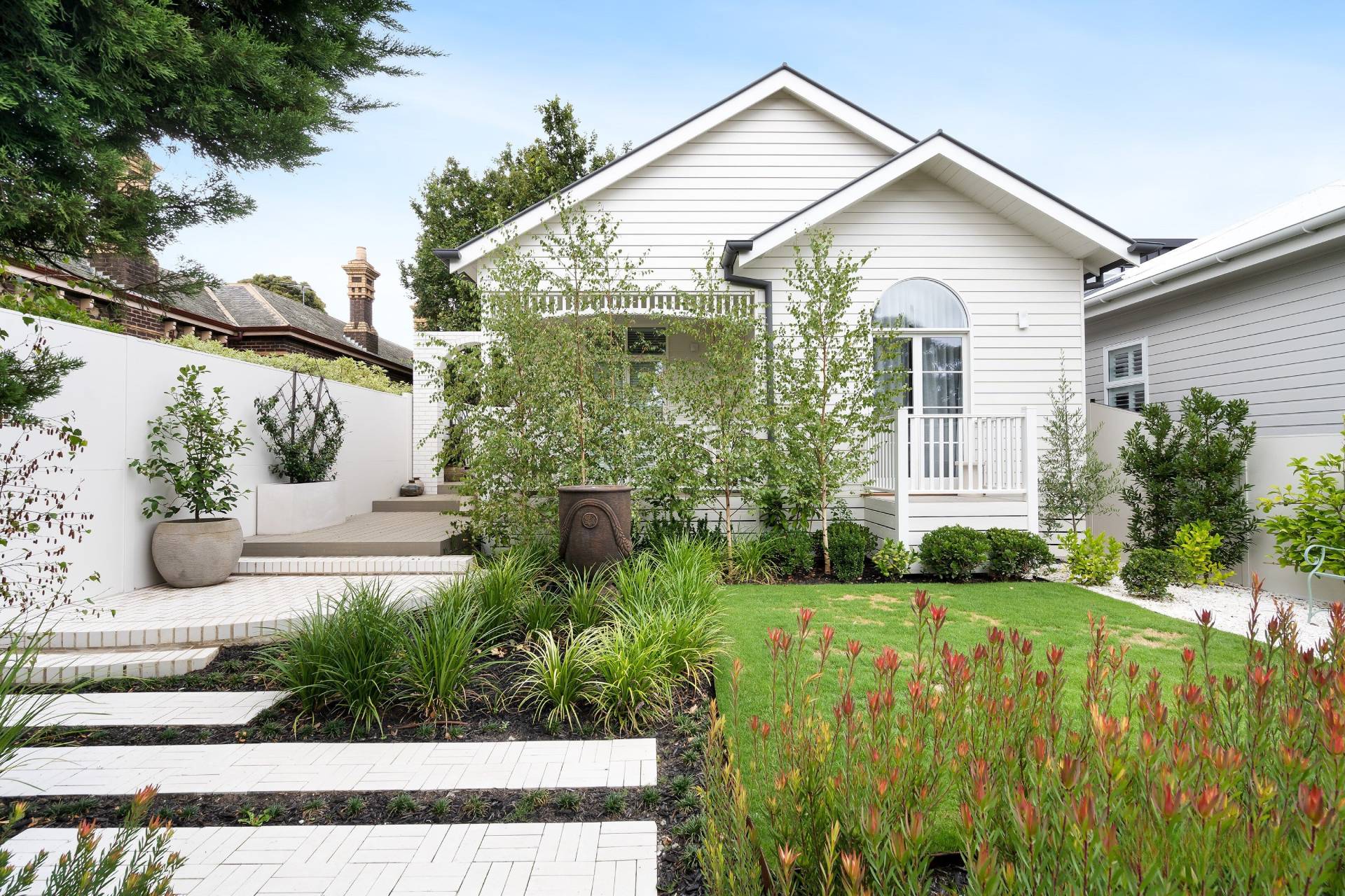 White house with manicured garden, green bushes, grass lawn, and a tiled walkway leading to the entrance.