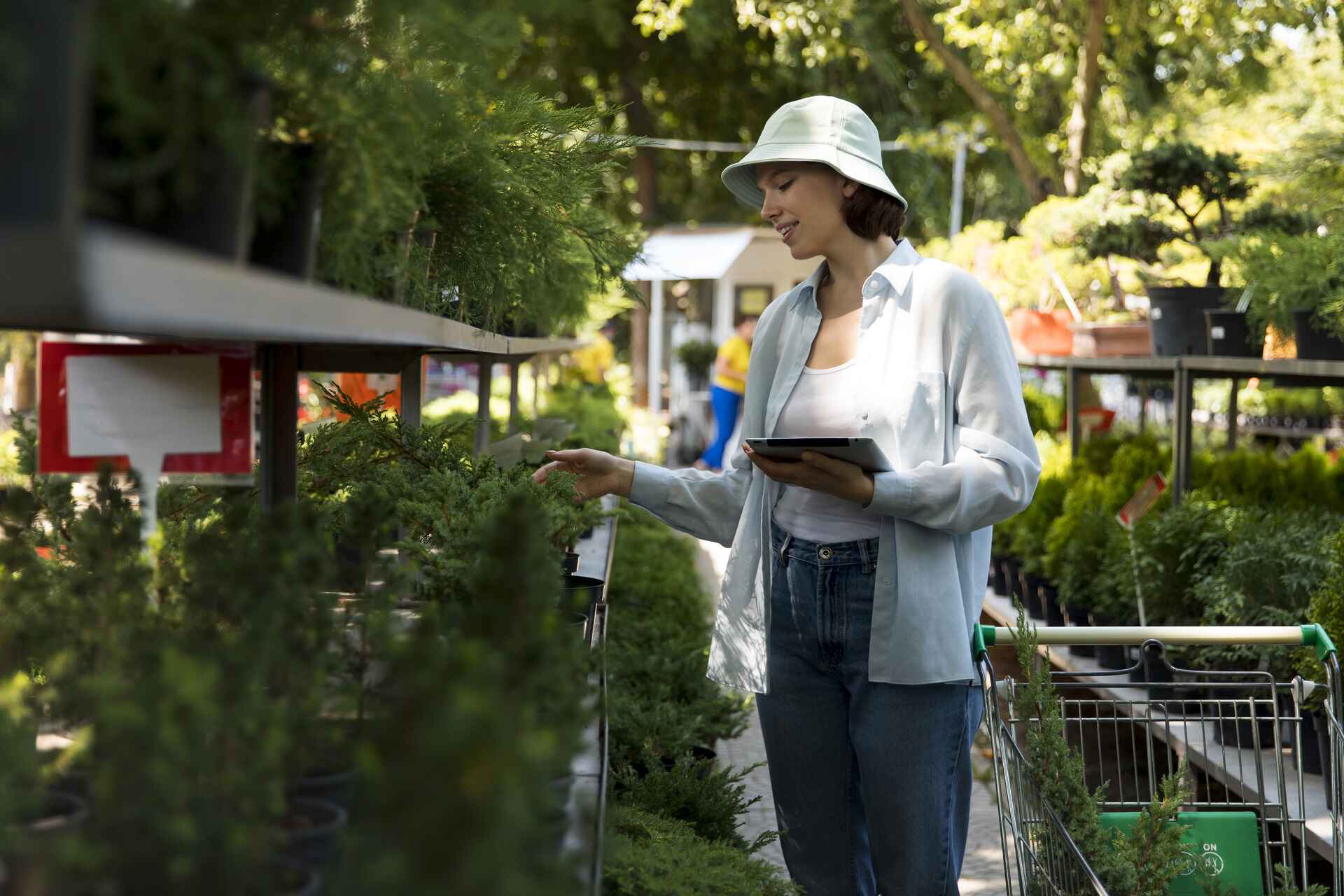 Woman shopping for plants at garden center, holding tablet and examining greenery