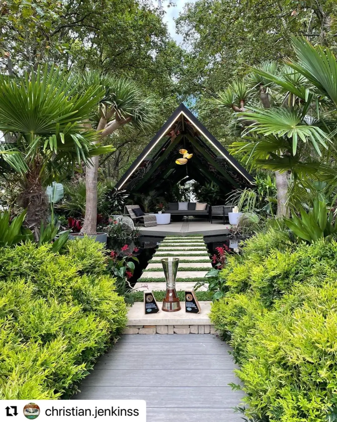 Tropical garden with a modern triangular pavilion, lush greenery, and a walkway leading to displayed trophies.