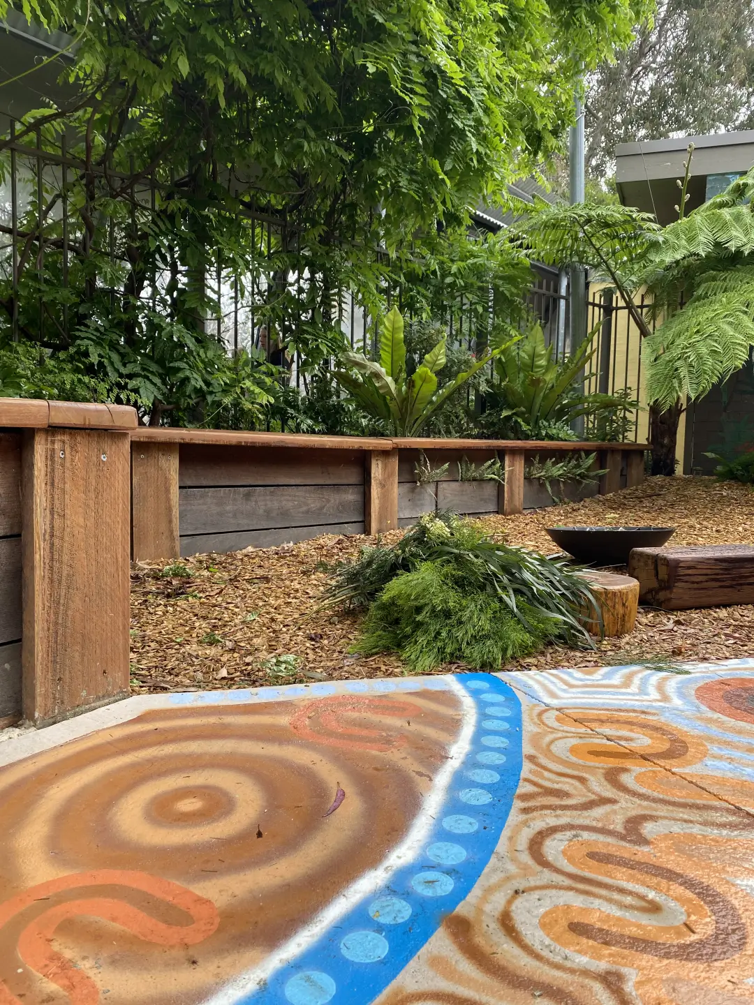 Garden area with leafy plants, wooden edging, and a path featuring Aboriginal-style artwork in earthy tones and blue accents.
