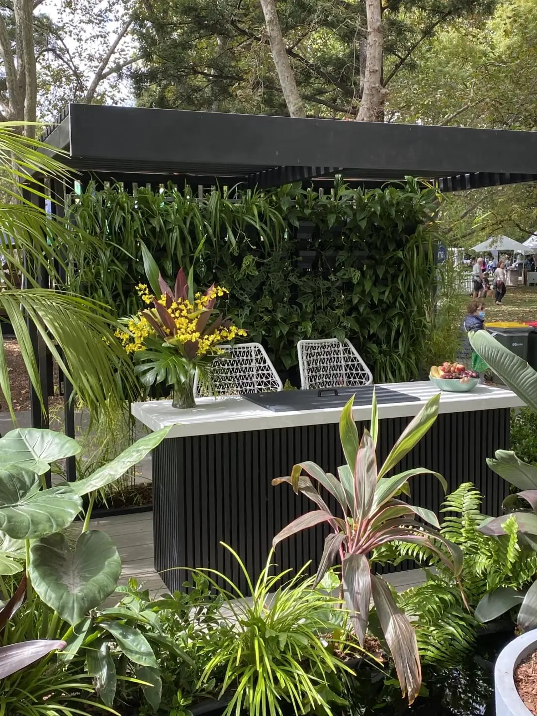 Modern garden display with lush plants, white chairs, and black pergola structure