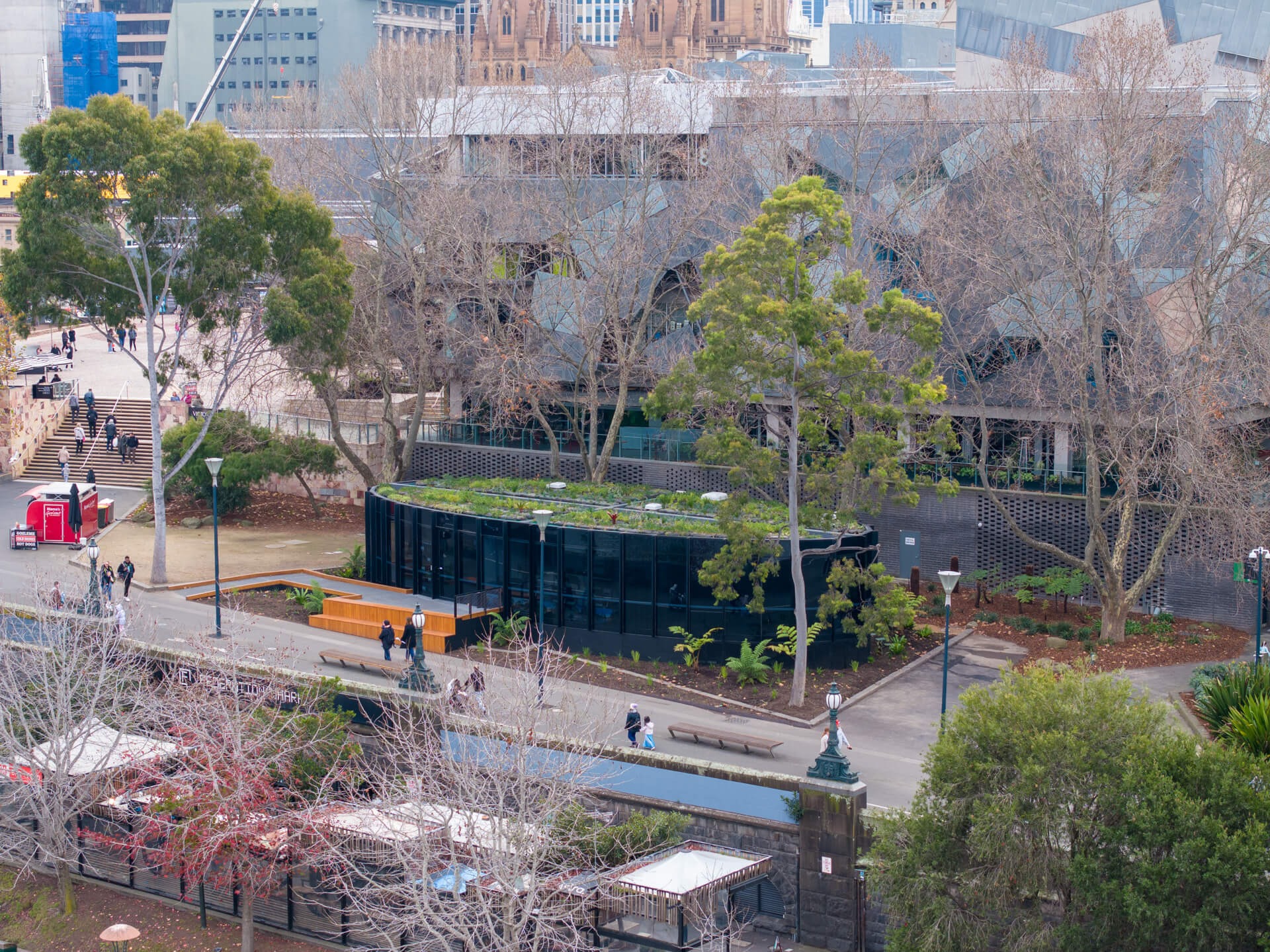 A low, black pavilion with a green roof sits among tall trees and walking paths in an urban park, with people strolling and city buildings in the background.