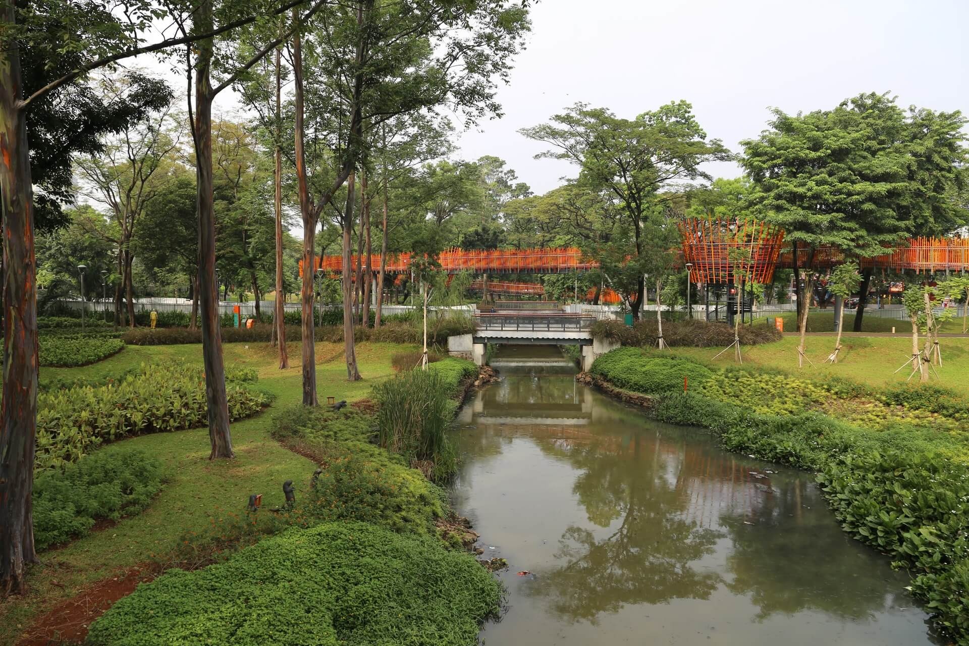 Tree-filled urban park with a narrow river, a small concrete bridge, and an orange elevated walkway in the background.