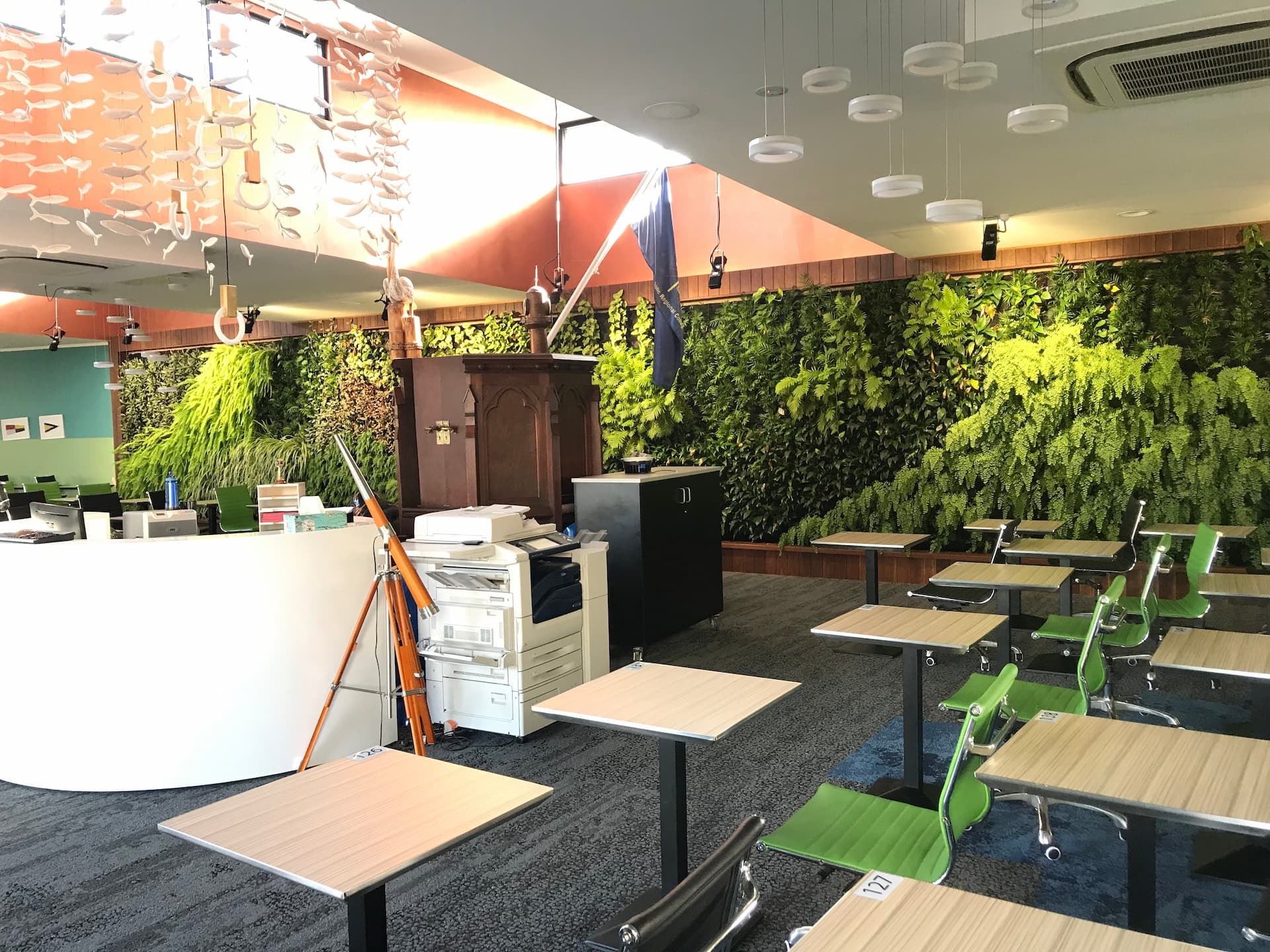 Bright open-plan office with desks, equipment and a long plant-covered living wall, showing how biophilic design brings greenery into the workplace.