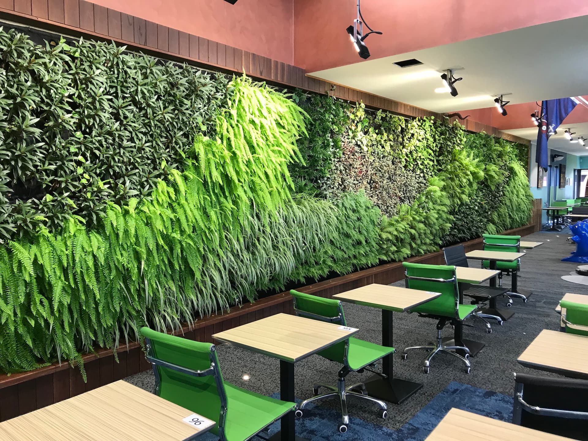 Indoor workspace with rows of desks and green chairs facing a large living wall of lush plants, illustrating biophilia in office design.