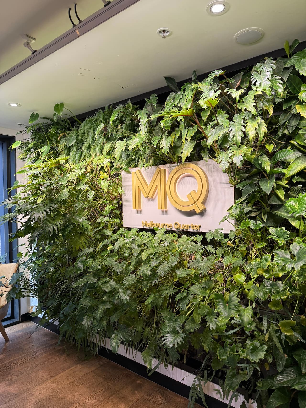 A dense green living wall with lush foliage surrounding a gold "MQ Melbourne Quarter" sign in a modern interior space.