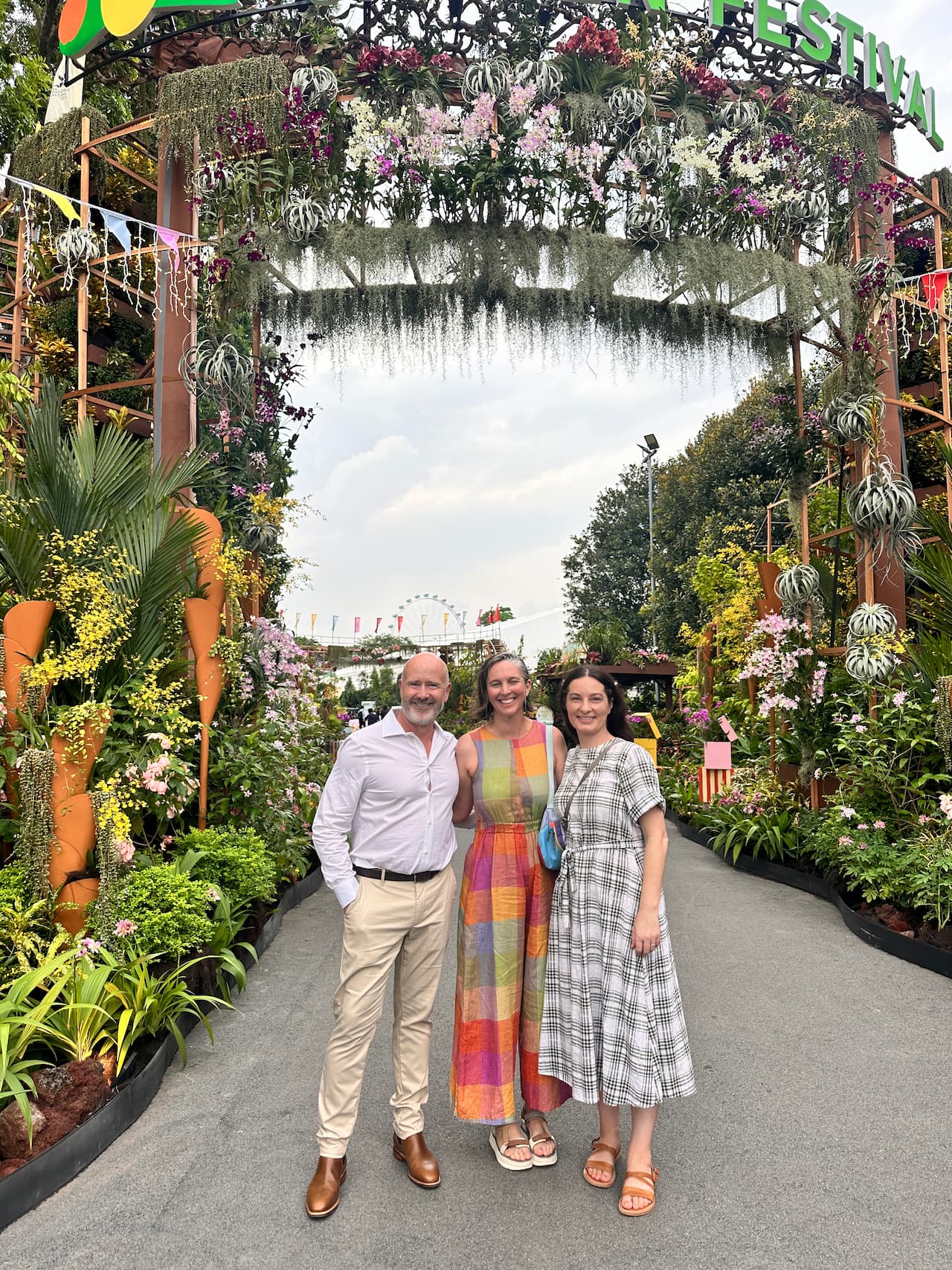 Michael Casey, Imogen Pullar, and Marnie Dawson standing on a garden path under a large floral archway decorated with hanging orchids and Spanish moss, with a Ferris wheel visible in the background at the Singapore Garden Festival.