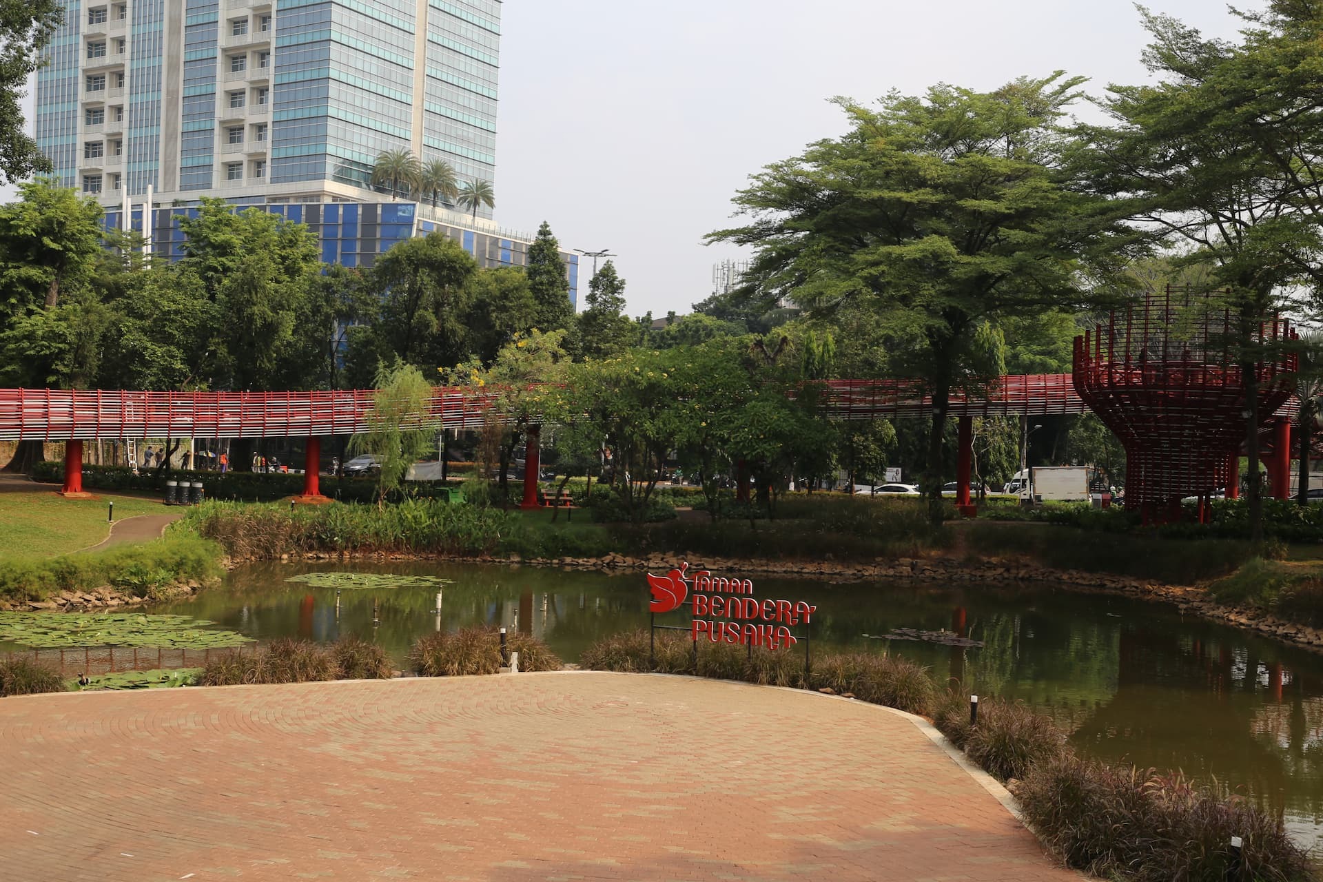 Taman Bendera Pusaka in Jakarta, showing a planted water body and red elevated walkway against nearby high-rise buildings.
