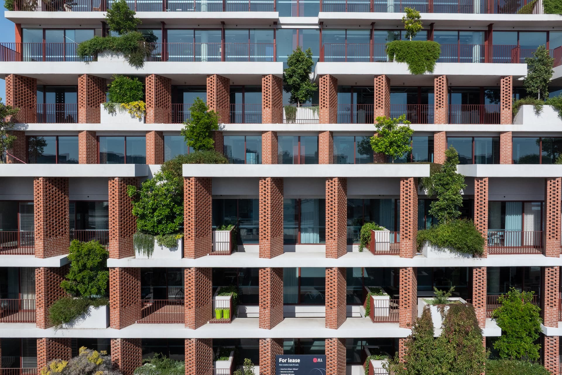 Façade greening on the Bruce St apartments: mature trees and trailing shrubs in built-in concrete planters on every level of a red brick and glass building.