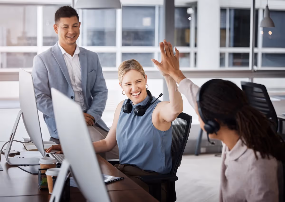 Three office workers, one celebrating with a high five, at desks with computers, showing teamwork and joy in a modern office setting.
