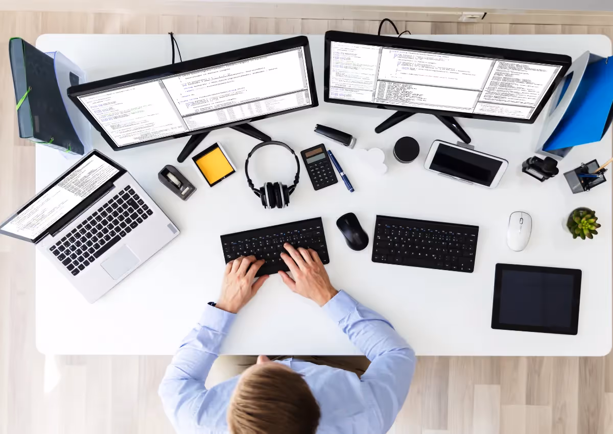 Top view of a workspace with a person using multiple screens and devices, showing various programs, alongside other tech accessories on a neat desk.