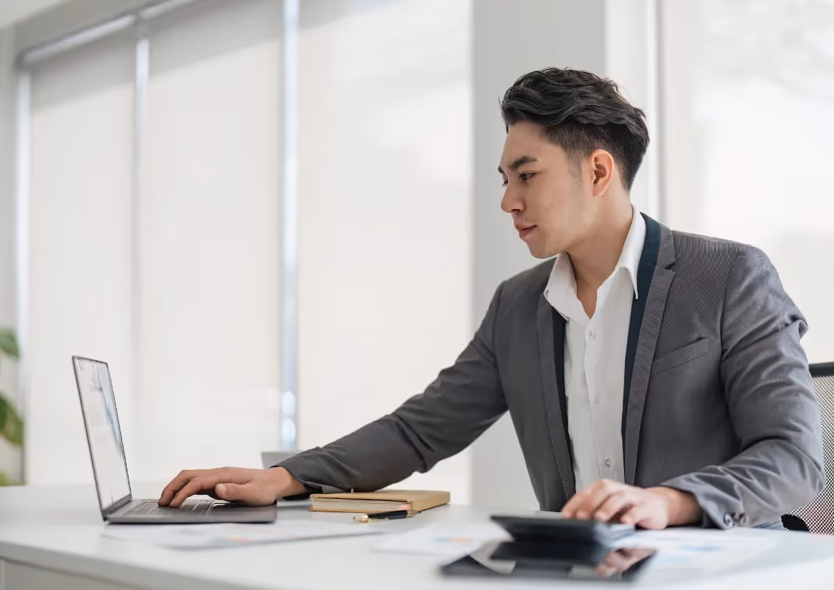 A businessman in a gray suit working intently at his desk, using a laptop and holding documents, in a bright office with large windows.