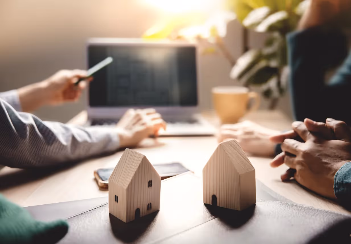 A close-up of a business meeting with three people around a table, featuring a laptop displaying blueprints and two small wooden house models, symbolizing a real estate or architectural discussion.