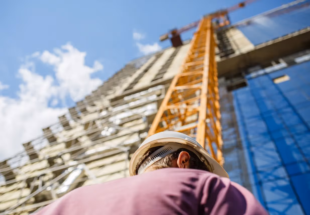 A construction worker wearing a hard hat looks up at a tall crane alongside a building under construction against a clear blue sky.