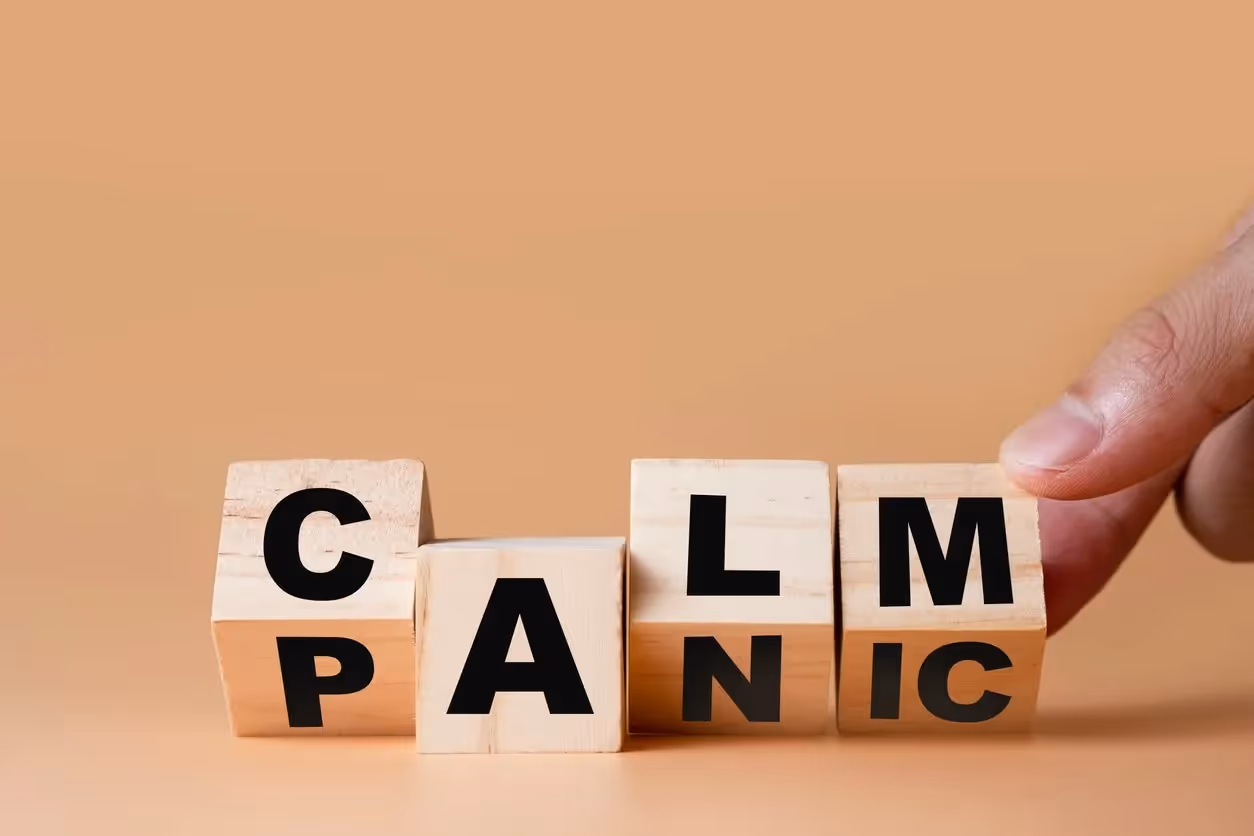 A hand flipping wooden blocks to change the word "panic" to "calm" on a plain beige background.