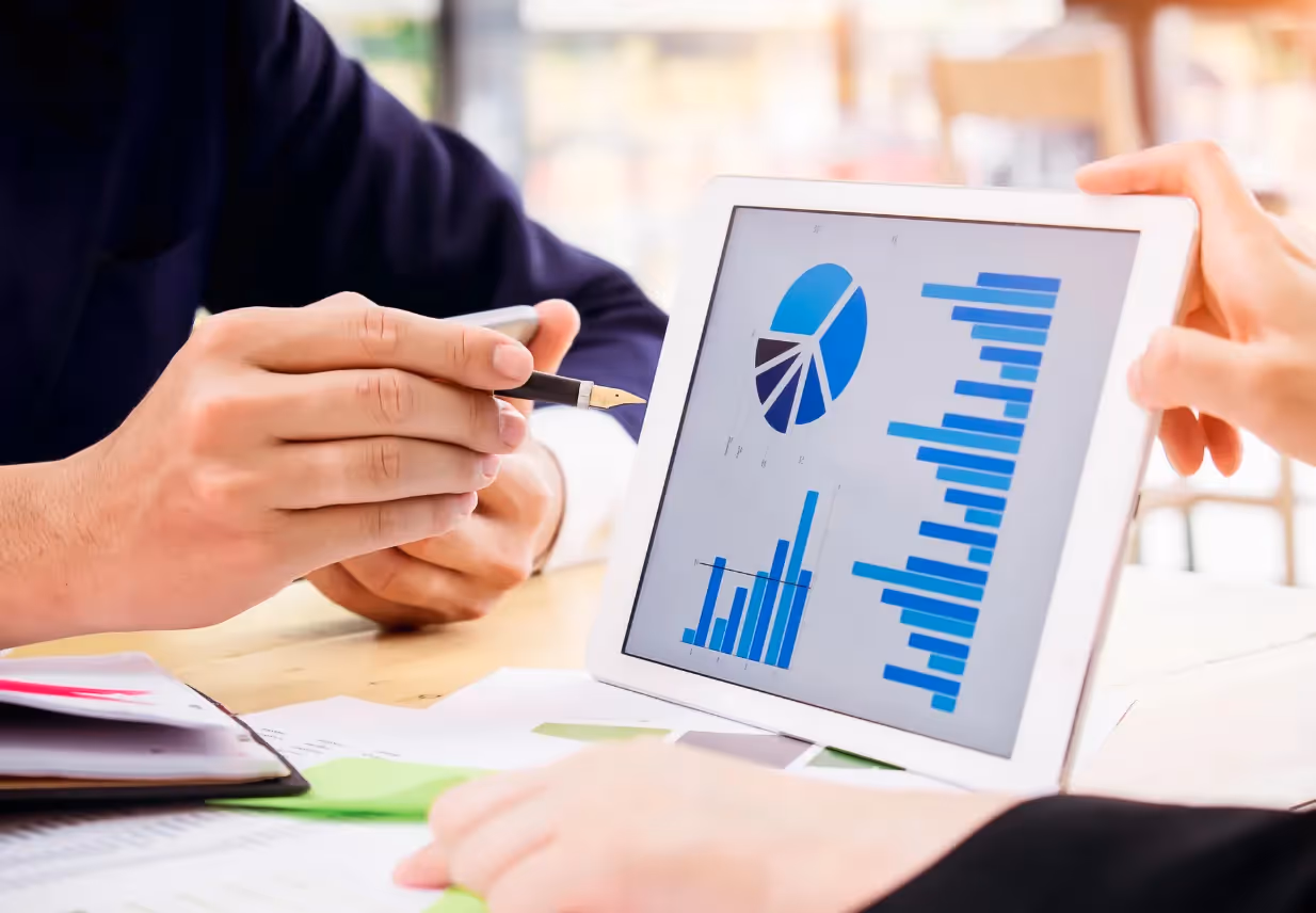 Two professionals review financial graphs on a tablet, surrounded by documents and a smartphone on the table.