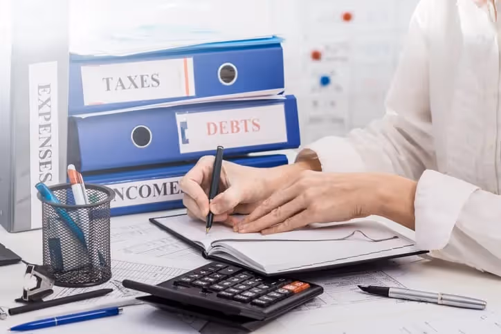A person writing in a notebook with a calculator on their desk surrounded by labeled binders including "taxes" and "income," indicating accounting tasks.