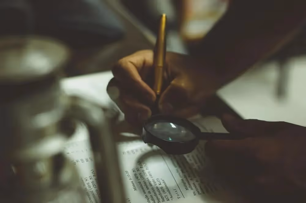 Close-up of hands holding a magnifying glass and a pencil over a document, suggesting detailed examination or study.
