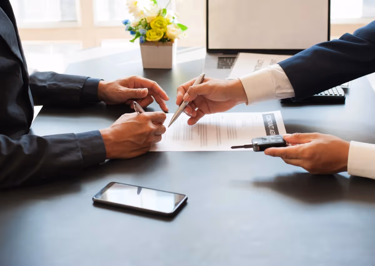 Two people in formal attire sit at a desk, one handing a pen to the other to sign a document. A smartphone and car keys are visible on the table.
