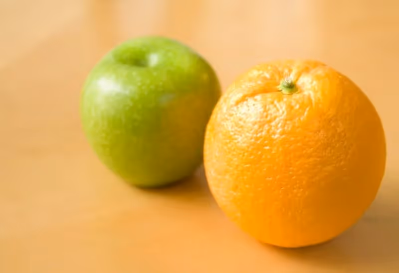 A green apple and an orange rest side by side on a light beige surface, highlighting their contrasting colors and textures.