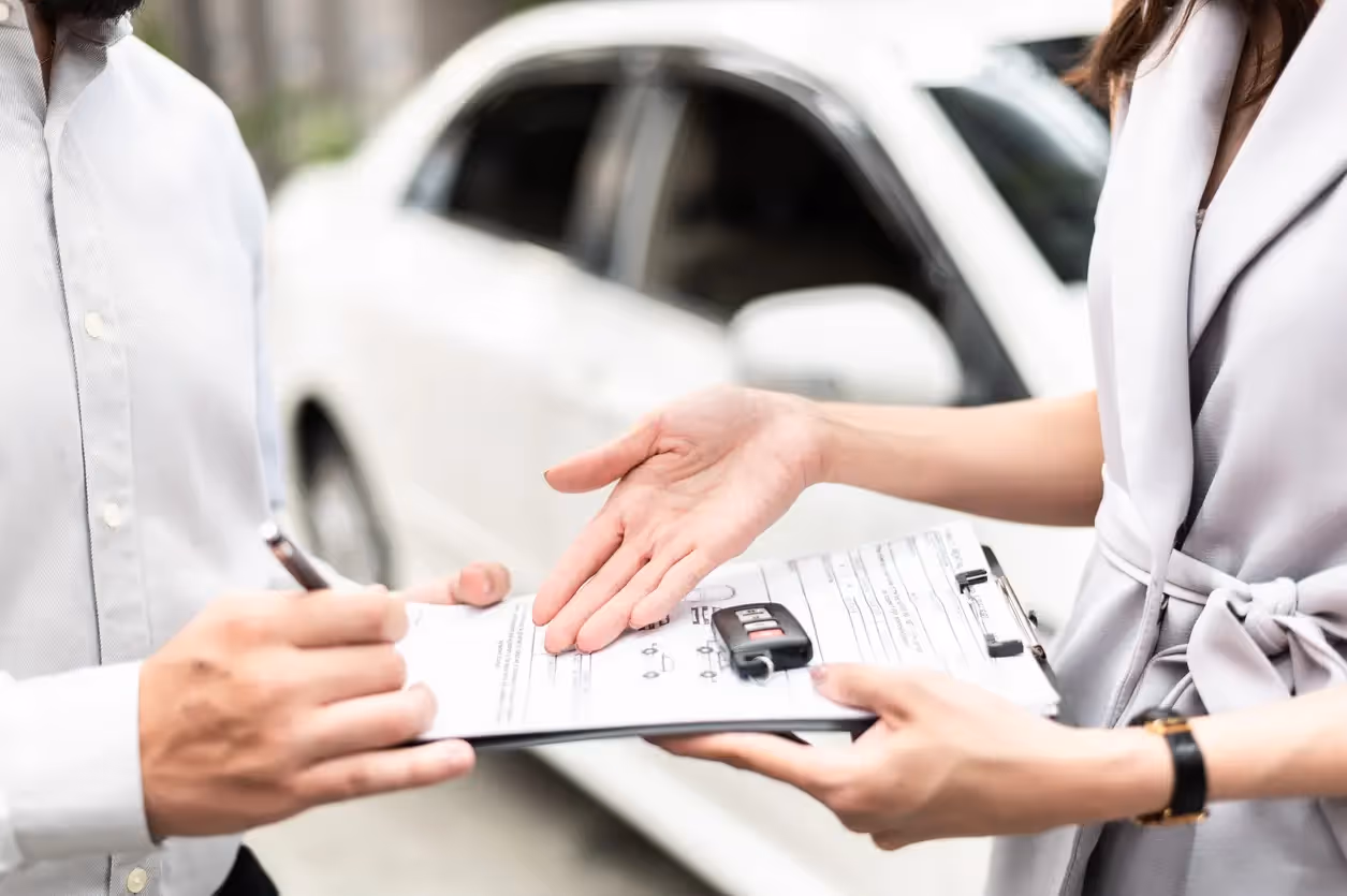 A close-up of a two people exchanging a document over a clipboard, with a white car in the background. The focus is on their hands and the document.