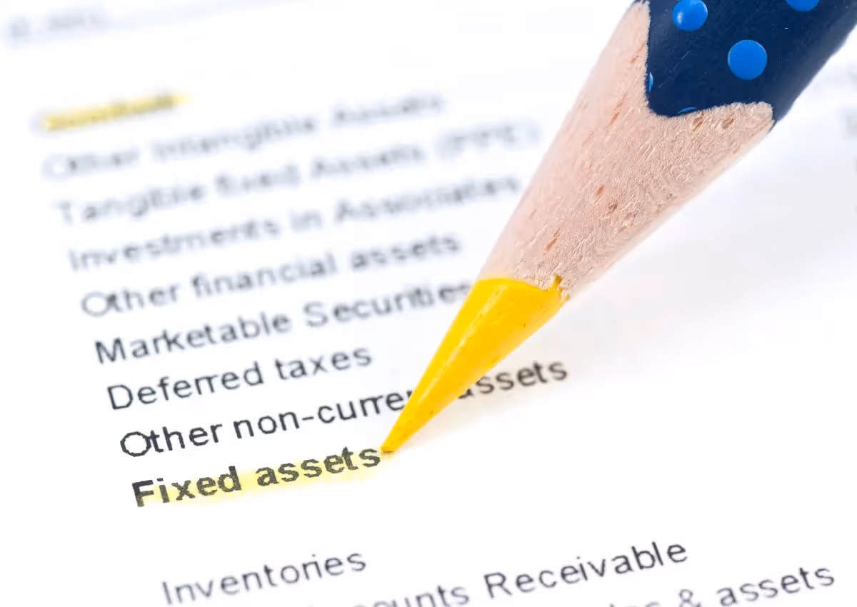A close-up of a pencil highlighting the phrase "Fixed assets" on a printed list. The paper includes various financial terms, such as "Tangible fixed assets" and "Deferred taxes," emphasizing financial documentation or accounting.