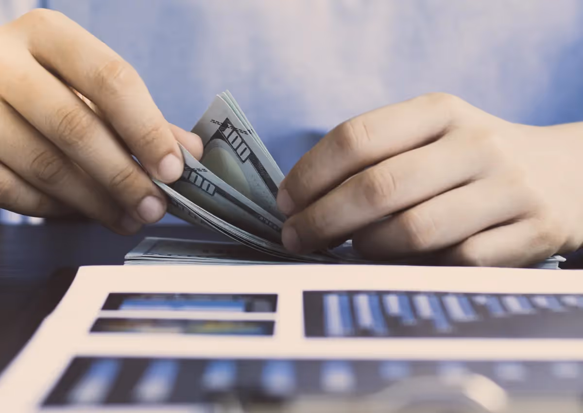 A person counting a stack of hundred-dollar bills with both hands, seated at a table. In the foreground, there's a blurred image of an open magazine or brochure. The person is wearing a light blue shirt.