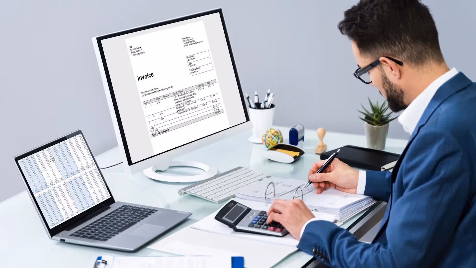 A person wearing glasses and a blue suit is sitting at a desk, using a calculator and writing on documents. There is a desktop monitor and a laptop displaying spreadsheets and invoices. The desk is organized with office supplies and plants.