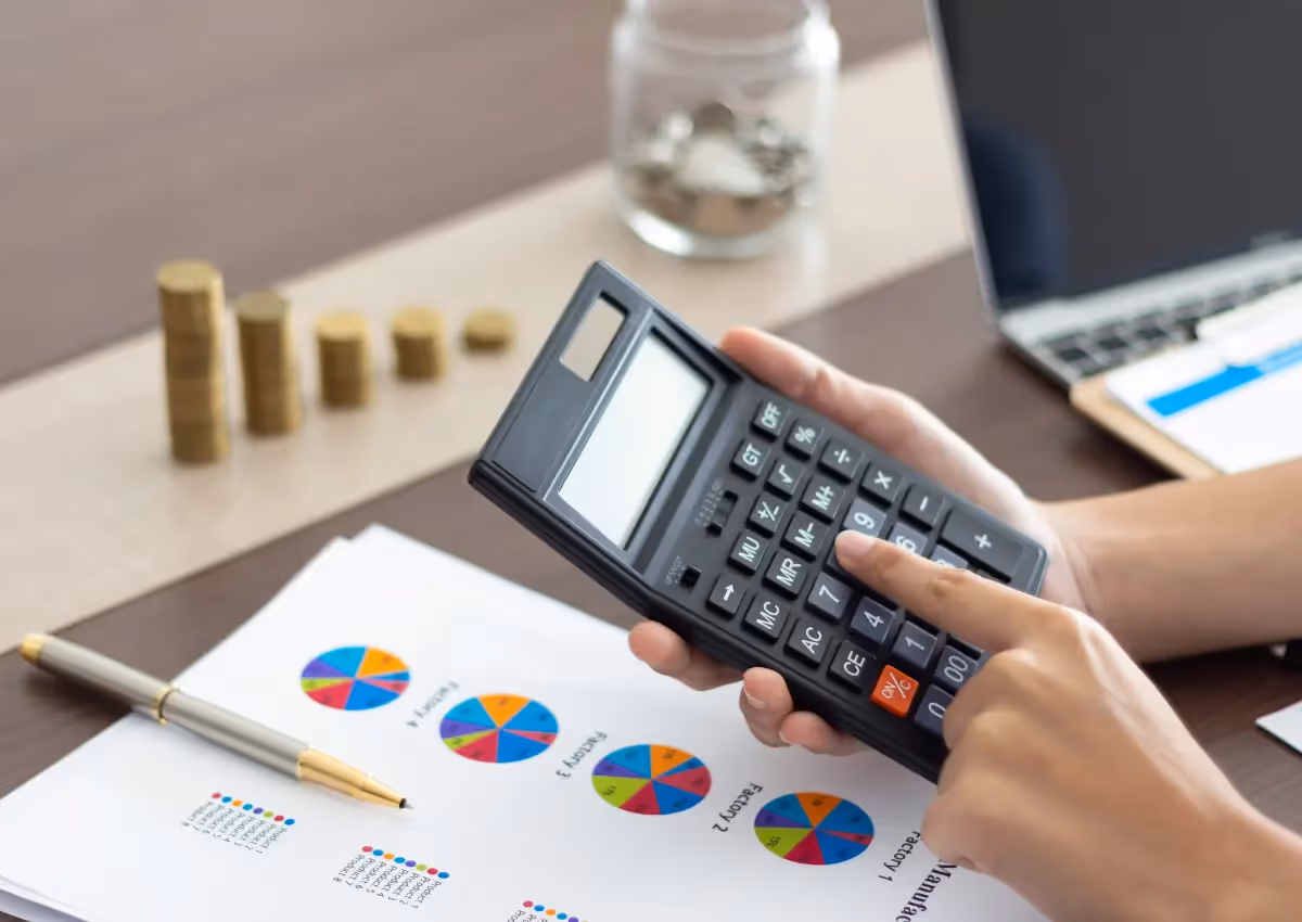 Person using a calculator on a desk with financial documents, charts, and pie graphs. A laptop, a glass jar with coins, and stacks of coins are in the background, alongside a pen.