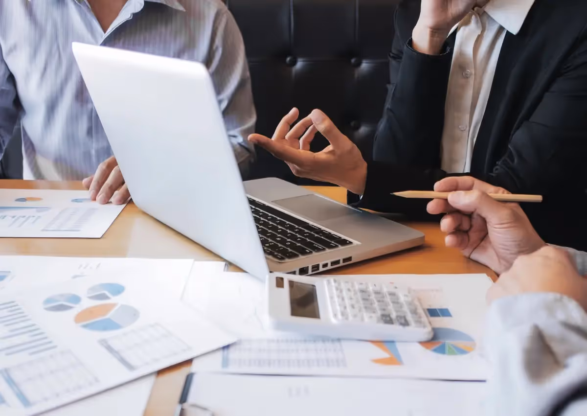 Three people are seated at a table with a laptop, documents featuring graphs, and a calculator. One person gestures with their hand while another holds a pencil, suggesting a discussion or meeting.
