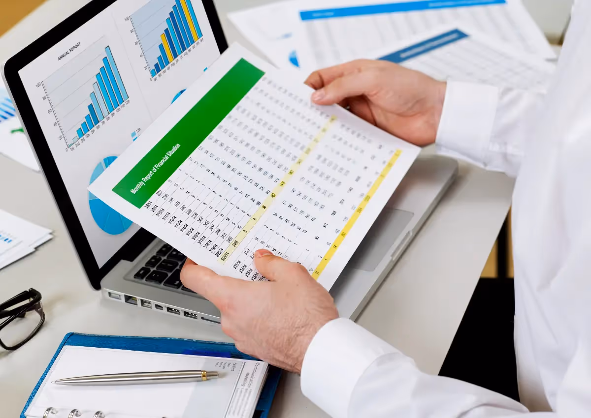 A person holds a printed spreadsheet filled with numerical data, analyzing it in front of a laptop displaying bar and pie charts. The desk also has a clipboard with papers and a pen. The setting indicates a focus on data analysis or financial evaluation.