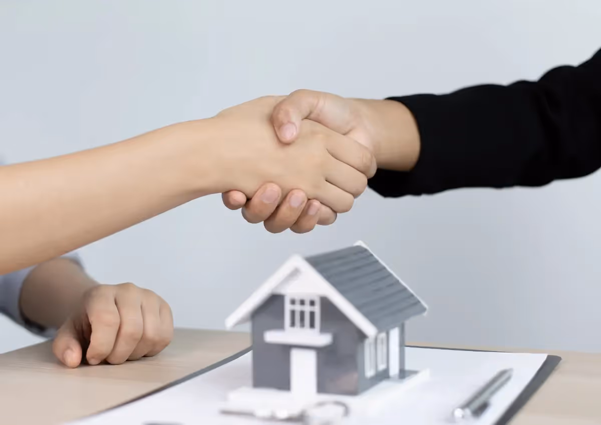 Two people shake hands over a table with a small house model, contract, and pen, symbolizing an agreement.
