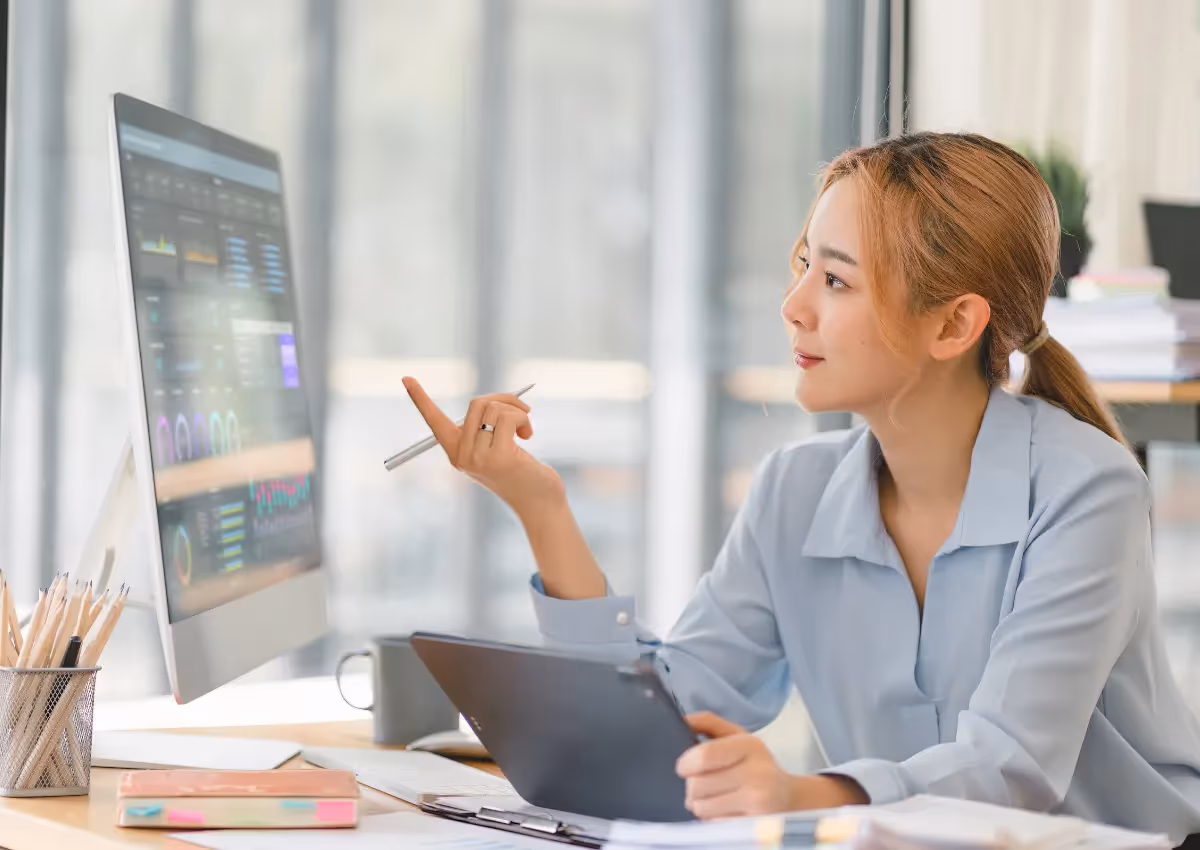 A woman in a light blue blouse sits at a desk, pointing at a computer screen displaying graphs and data. She holds a stylus in her other hand and has a focused expression. The office environment is bright and modern.