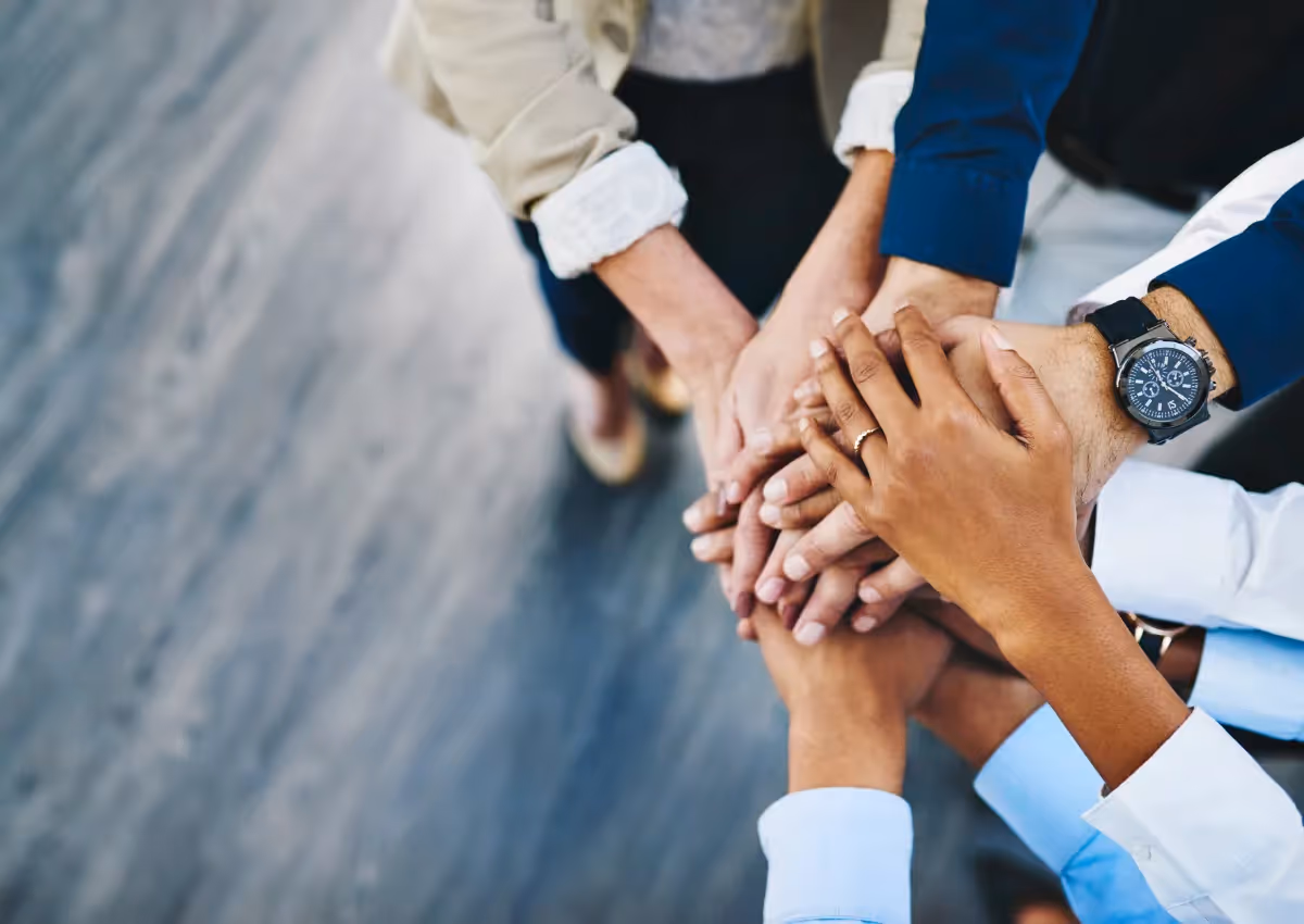 A close-up of a diverse group of people standing in a circle, stacking their hands together in a show of unity and teamwork. The background is a blurred gray surface.