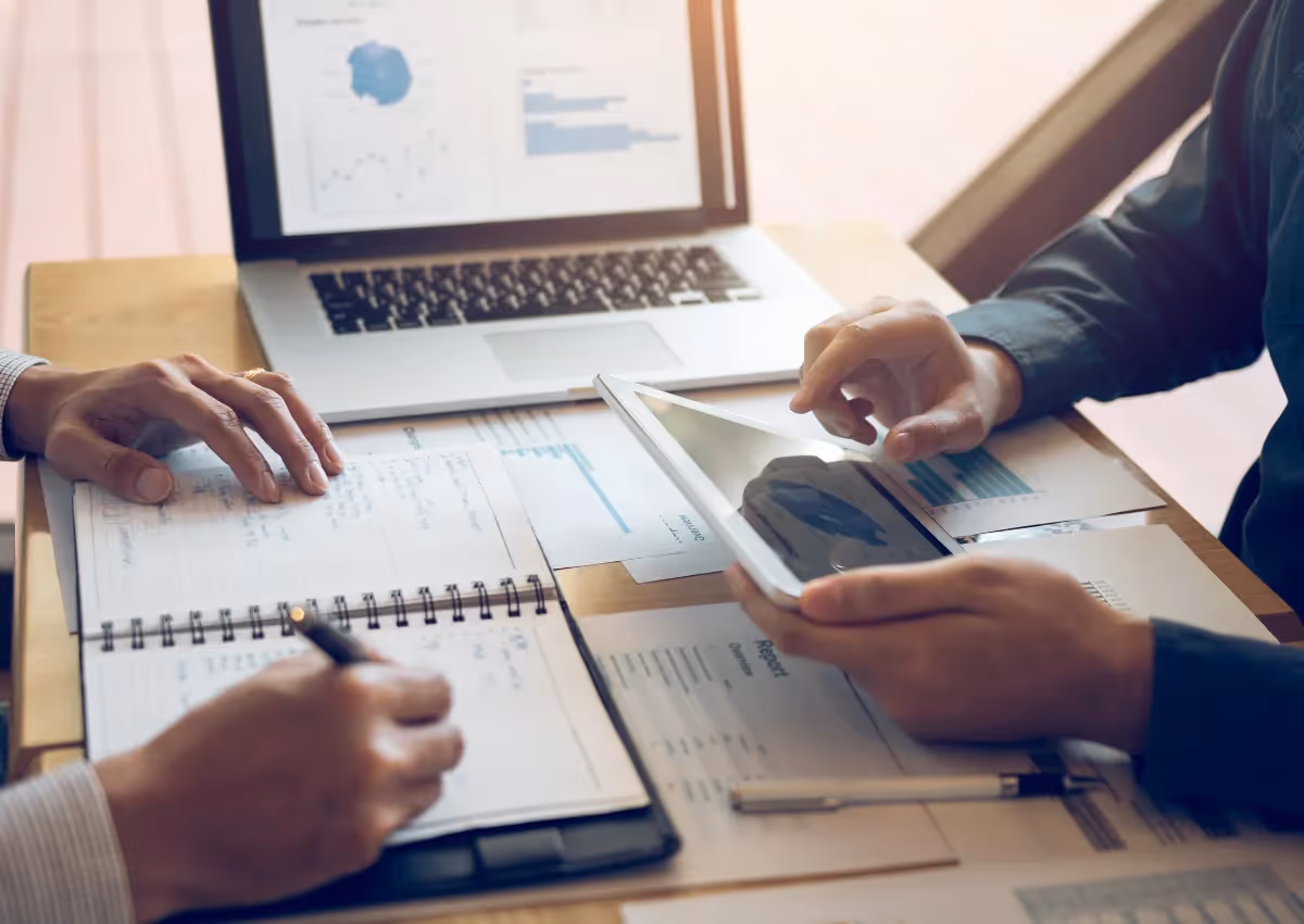 Two people collaborate at a table with a laptop, tablet, and documents. One person takes notes in a notebook while the other gestures at the tablet. The laptop screen displays charts and graphs. The scene is well-lit, suggesting a work or study environment.