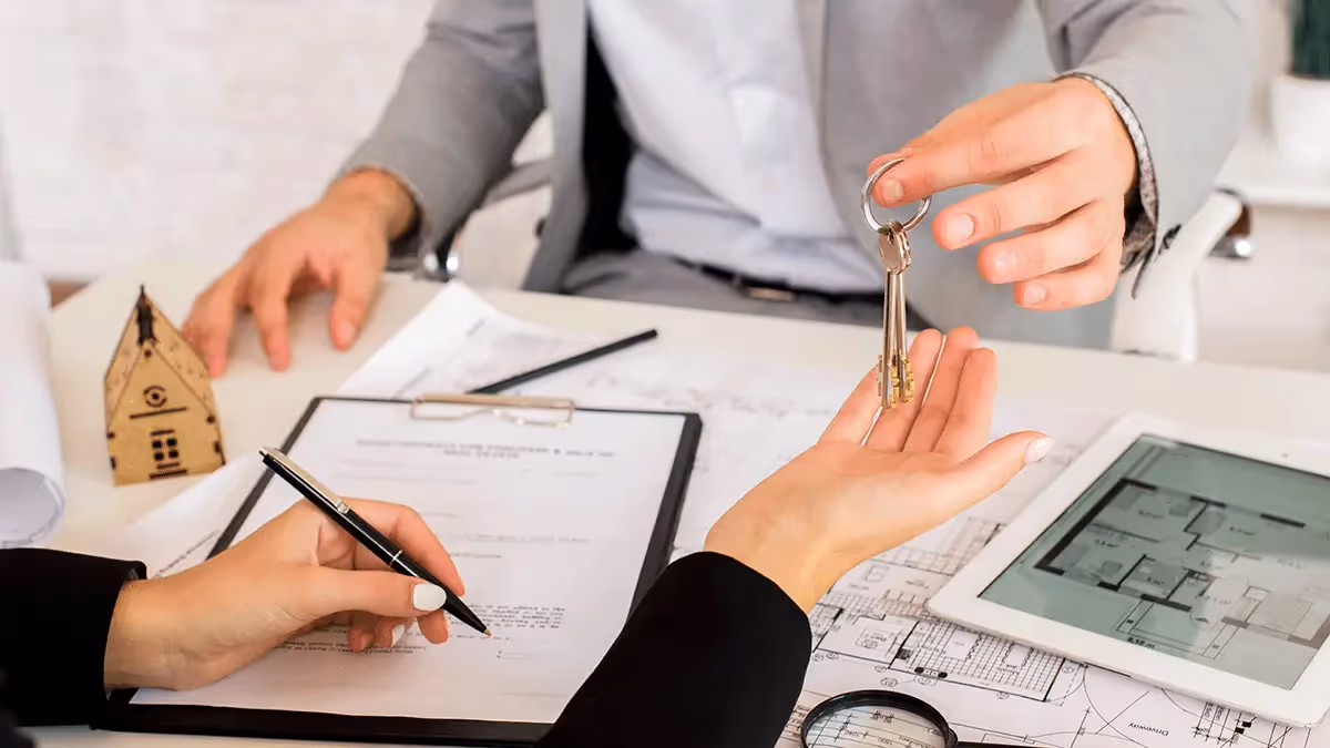 A person hands keys to a client across a table with house plans, a model home, and documents, symbolizing a transaction.