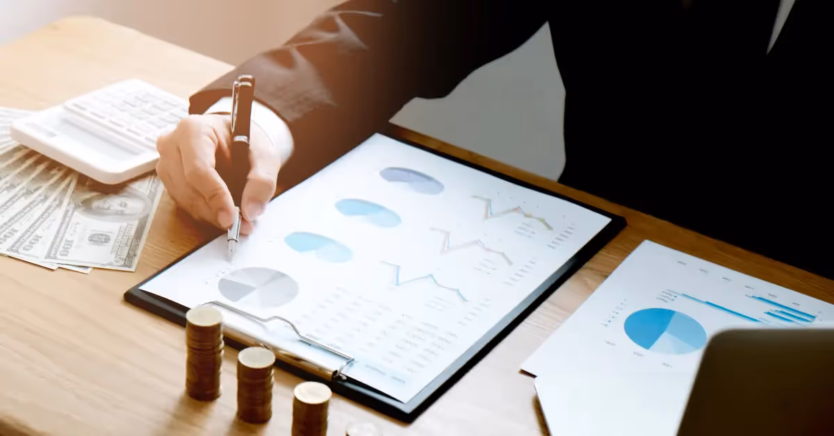A person in formal attire uses a pen to point at a clipboard with financial charts. A calculator, stacks of coins, and US dollar bills are on the desk, suggesting financial planning or analysis.