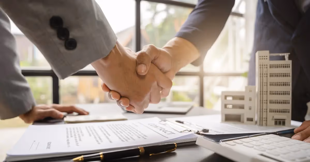 Two people shaking hands over a table with a lease contract and building mock-ups