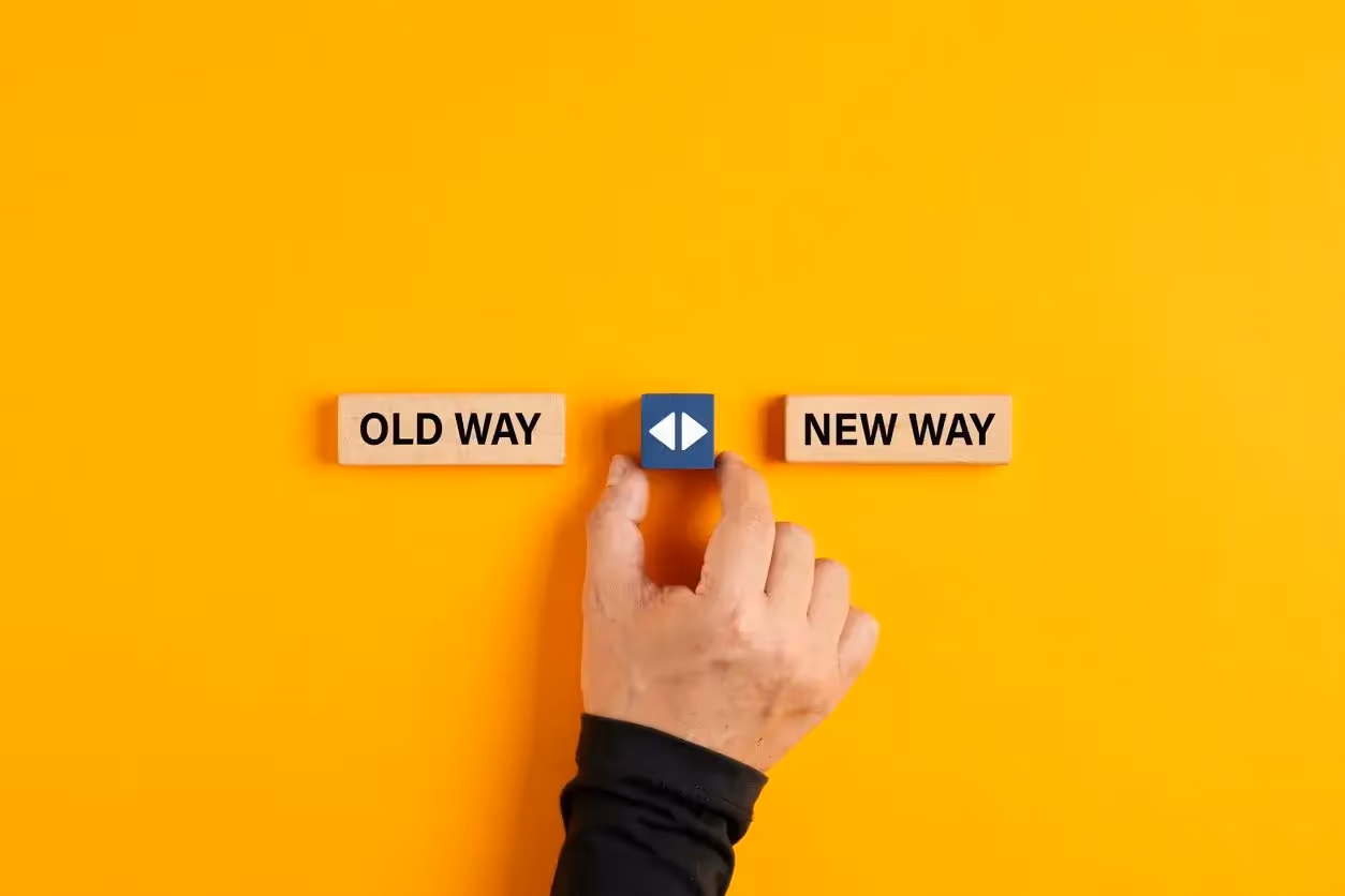 A person's hand pressing a blue button positioned between two wooden blocks. the left block reads "old way" and the right reads "new way," against a bright orange background.