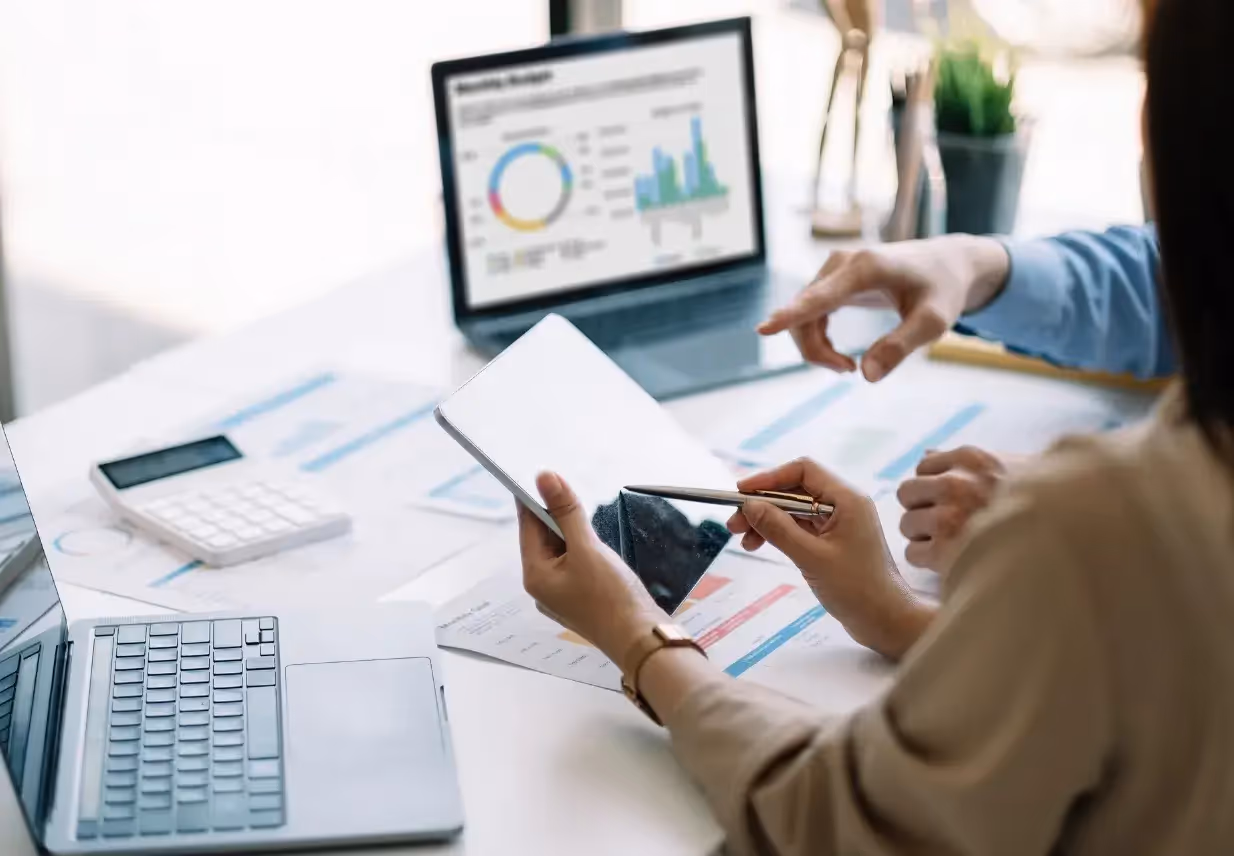 Two professionals analyze data on printed charts and a laptop displaying graphs in a bright office setting, with calculators and digital devices on the table.
