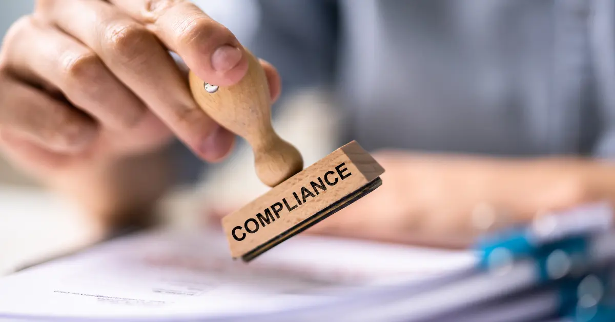 A hand holds a rubber stamp reading compliance over a stack of financial documents.