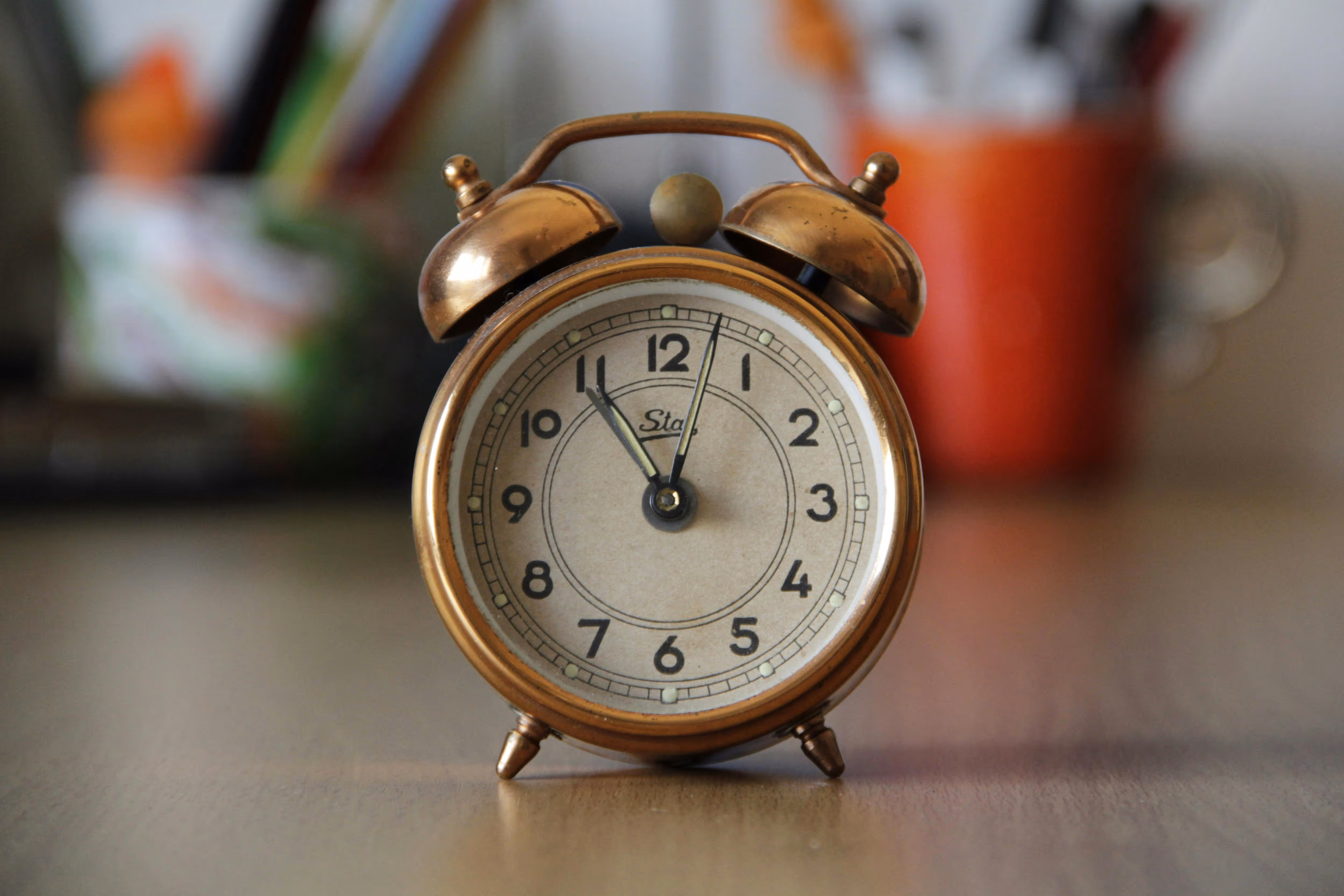 Close-up of a vintage brass alarm clock showing 11:03 on a wooden surface with blurred background objects.