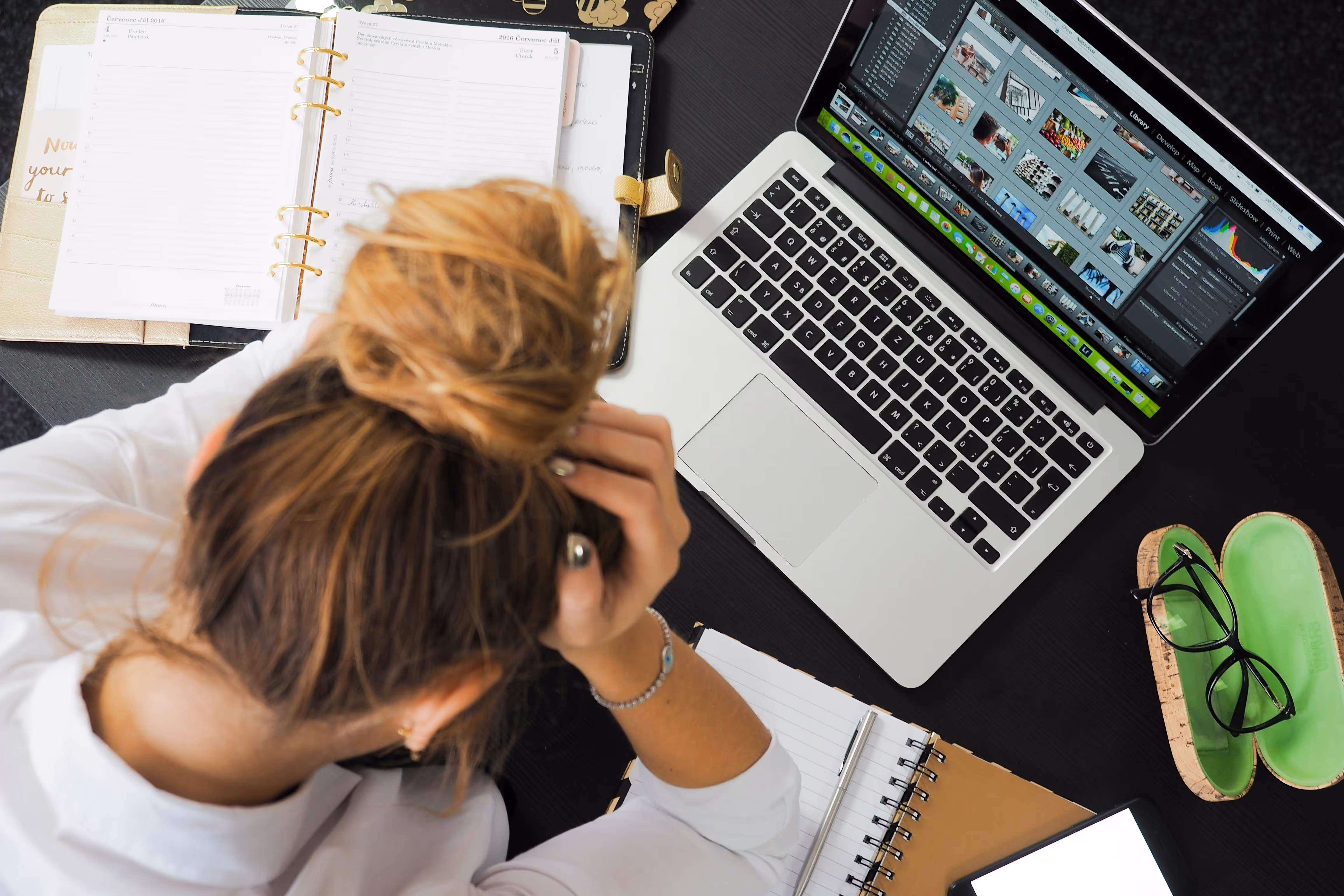 Person with hair in a bun holding their head at a desk with a laptop, open planner, notebook, tablet, and glasses case.