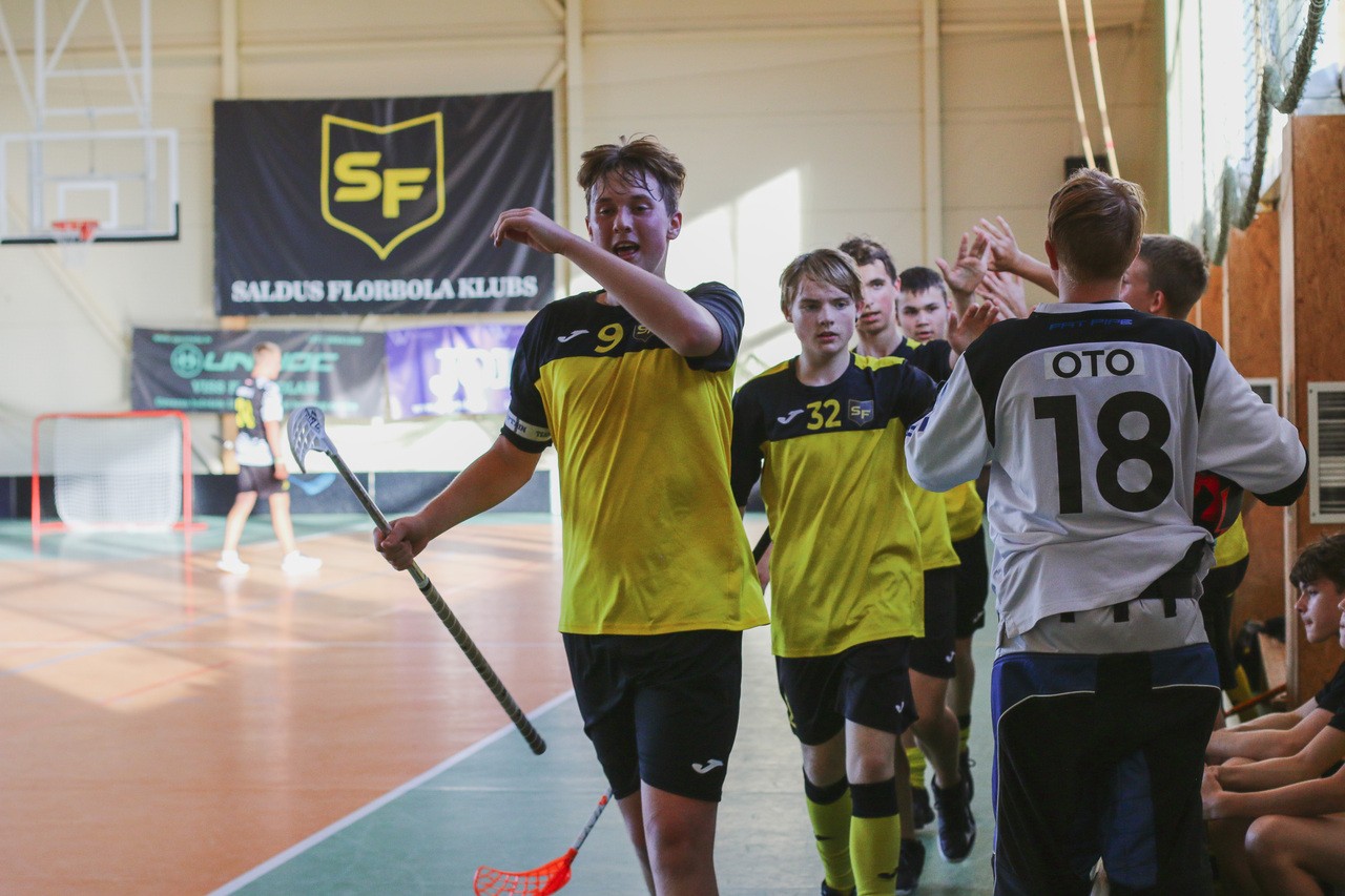 Florbola spēlētāji dzeltenās un melnās formās sveicinās viens otru sporta zālē pie vārtiem.