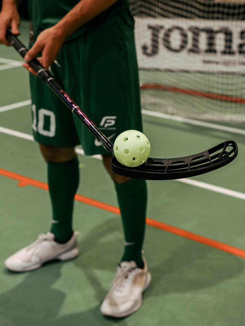 Florbola spēlētājs zaļā formā ar numuru 20, turot nūju ar perforētu bumbiņu uz tās sporta laukumā.