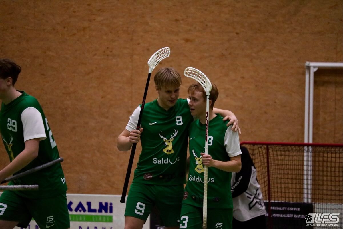 Divi florbola spēlētāji zaļās formās apskauj viens otru iekš telpu laukuma pie vārtiem.