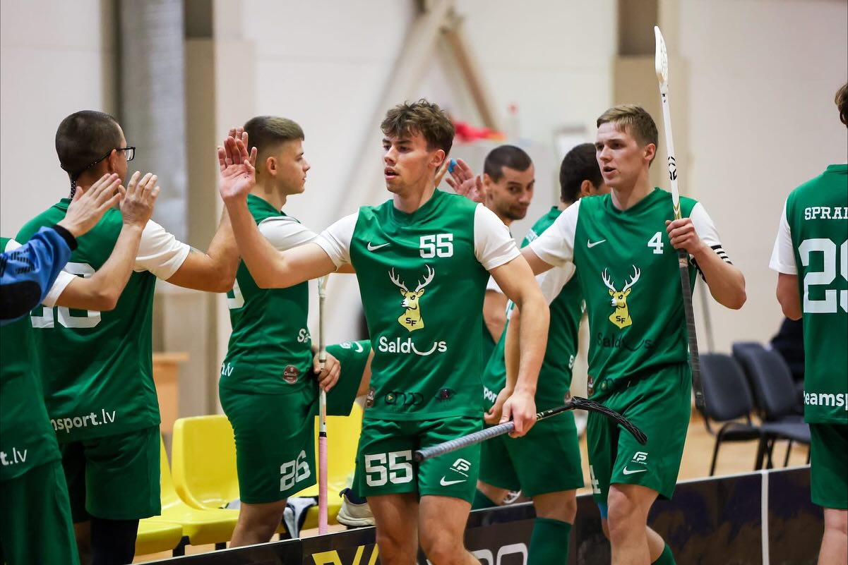 Florbola spēlētāji zaļos formas tērpos ar nūjām sveic viens otru ar augšupejošiem plaukstu sitieniem sporta zālē.